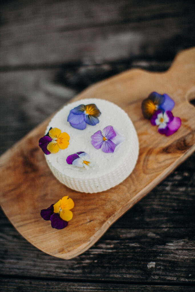 Runder Frischkäse dekoriert mit essbaren Blüten auf Holzbrett – Biohof Pamer